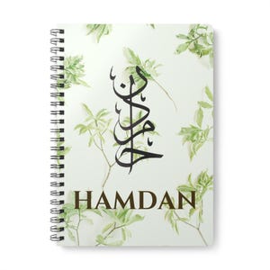 Puede incluir: Cuaderno de espiral con una cubierta blanca que presenta un diseño de caligrafía negra y el nombre "HAMDAN" en marrón. La cubierta está decorada con ramas de hojas verdes, creando una estética natural.