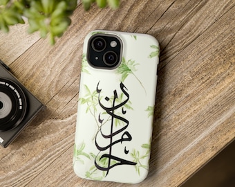 May include: White phone case with a green floral pattern and black Arabic calligraphy. The case is on a wooden surface with a camera and greenery in the background. The phone case is designed to protect the phone from scratches and damage.