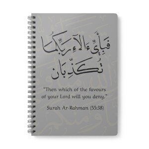 May include: A spiral-bound notebook with a gray cover featuring Arabic calligraphy and English text. The text reads, "Then which of the favours of your Lord will you deny." and "Surah Ar-Rahman (55:38)." The notebook has a silver spiral binding.