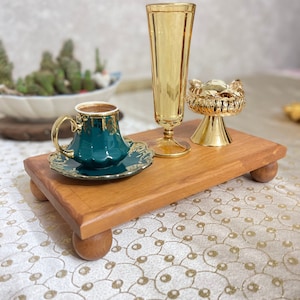 Könnte beinhalten: Ein Holztablett mit abgerundeten Beinen, auf dem eine grüne und goldene Kaffeetasse mit Untertasse, ein goldener Dekorständer und ein hohes, klares Glas mit goldfarbenem Rand stehen.