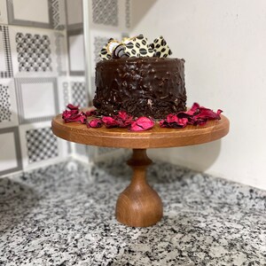 Puede incluir: Un pastel de chocolate con virutas de chocolate y decoraciones de chocolate blanco en la parte superior, sentado en un soporte para pasteles de madera con pétalos de flores rosas.
