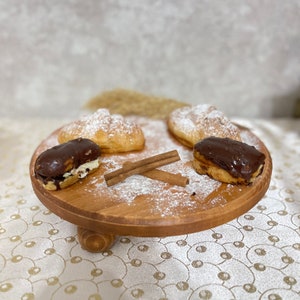 Puede incluir: Un soporte para pasteles de madera con cuatro pasteles encima. Dos de los pasteles están rellenos de crema y cubiertos con glaseado de chocolate. Los otros dos pasteles están rellenos de crema y espolvoreados con azúcar en polvo. Hay dos palitos de canela en el soporte.
