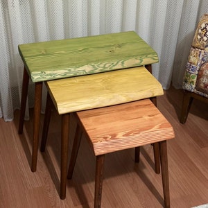 Puede incluir: Tres mesas nido de madera con acabado de veta de madera natural. La mesa superior está pintada de verde, la mesa del medio está pintada de amarillo y la mesa inferior está pintada de naranja. Las mesas tienen patas de color marrón oscuro.