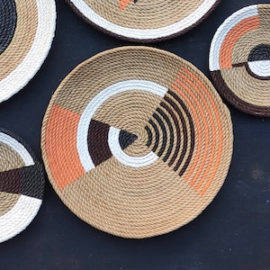 Puede incluir: Colecci&oacute;n de platos decorativos de cuerda tejida con dise&ntilde;os geom&eacute;tricos. Los platos presentan un color beige natural con acentos de blanco, marr&oacute;n, negro y naranja. Los platos est&aacute;n dispuestos sobre una superficie oscura.