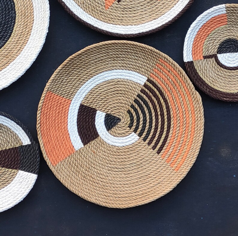 Puede incluir: Colecci&oacute;n de platos decorativos de cuerda tejida con dise&ntilde;os geom&eacute;tricos. Los platos presentan un color beige natural con acentos de blanco, marr&oacute;n, negro y naranja. Los platos est&aacute;n dispuestos sobre una superficie oscura.