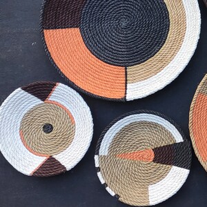 Puede incluir: Una colecci&oacute;n de cuencos decorativos de cuerda tejida en varios tama&ntilde;os y dise&ntilde;os geom&eacute;tricos. Los cuencos presentan una mezcla de colores, incluyendo negro, naranja, blanco, marr&oacute;n y beige. Los cuencos est&aacute;n dispuestos sobre una superficie oscura.