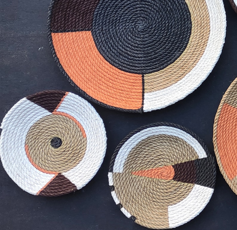 Puede incluir: Una colecci&oacute;n de cuencos decorativos de cuerda tejida en varios tama&ntilde;os y dise&ntilde;os geom&eacute;tricos. Los cuencos presentan una mezcla de colores, incluyendo negro, naranja, blanco, marr&oacute;n y beige. Los cuencos est&aacute;n dispuestos sobre una superficie oscura.