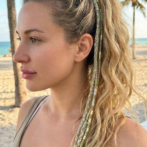 Op de afbeelding: Blond haar in een hoge paardenstaart met drie gevlochten haarversieringen. De versieringen hebben een mix van groene, bruine en gouden draden, wat een decoratief element aan het kapsel toevoegt. De achtergrond toont een strandscène met palmbomen.
