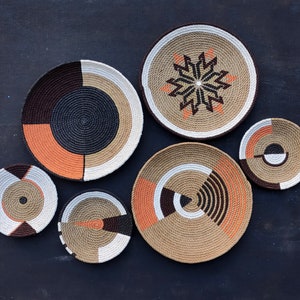Puede incluir: Una colecci&oacute;n de cuencos decorativos tejidos en varios tama&ntilde;os y dise&ntilde;os. Los cuencos presentan una mezcla de colores que incluyen marr&oacute;n, negro, naranja y blanco. Algunos cuencos tienen patrones geom&eacute;tricos, mientras que otros tienen dise&ntilde;os en forma de estrella. Los cuencos est&aacute;n dispuestos sobre una superficie oscura.