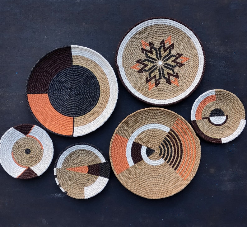 Puede incluir: Una colecci&oacute;n de cuencos decorativos tejidos en varios tama&ntilde;os y dise&ntilde;os. Los cuencos presentan una mezcla de colores que incluyen marr&oacute;n, negro, naranja y blanco. Algunos cuencos tienen patrones geom&eacute;tricos, mientras que otros tienen dise&ntilde;os en forma de estrella. Los cuencos est&aacute;n dispuestos sobre una superficie oscura.