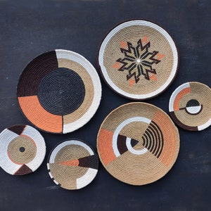 Puede incluir: Colecci&oacute;n de siete cestas de pared tejidas de varios tama&ntilde;os y dise&ntilde;os. Las cestas presentan una mezcla de colores naturales, marr&oacute;n, negro, blanco y naranja. Algunas tienen patrones geom&eacute;tricos, mientras que otras tienen dise&ntilde;os en forma de estrella o circulares.