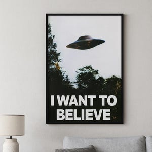 Può includere: Poster incorniciato nero con un'immagine di un UFO sopra una fila di alberi e la scritta "I WANT TO BELIEVE" in bianco. L'immagine ha un'estetica vintage, leggermente sfocata. Il poster è appeso a una parete bianca sopra un divano grigio.