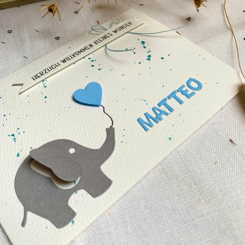 以下が含まれることがあります： グレーの象が、薄い青色のハート型の風船を持っています。風船には「HERZLICH WILLKOMMEN KLEINES WUNDER」という文字が書かれたバナーと、青い文字で「MATTEO」という名前が書かれています。
