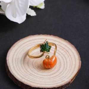 May include: A gold ring with a green leaf and an orange pumpkin charm. The ring is displayed on a wooden base.