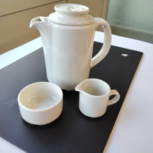 May include: A white ceramic coffee pot with a lid, a white ceramic creamer, and a white ceramic sugar bowl on a black surface.