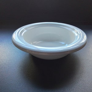 Puede incluir: Un tazón de cerámica blanco con un borde ligeramente elevado. El tazón está vacío y tiene un acabado brillante.