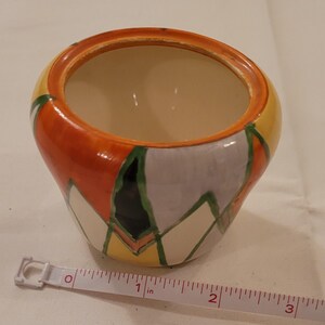 May include: A ceramic bowl with a cream-colored interior and a hand-painted exterior featuring orange, yellow, green, and lavender geometric shapes. A white measuring tape is positioned in front, showing the bowl is approximately 3 inches wide.