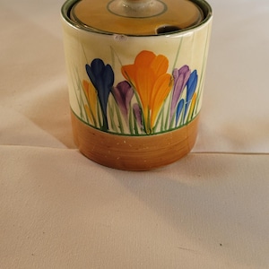 May include: A cylindrical ceramic sugar bowl with a lid featuring a hand-painted floral design of orange, purple, and blue crocus flowers on a cream background. The base and lid are accented with a solid brown band, and the lid has a small knob.