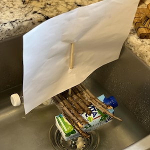 May include: A homemade boat made from a cardboard juice box, sticks, and a piece of paper for a sail. The boat is floating in a stainless steel kitchen sink.