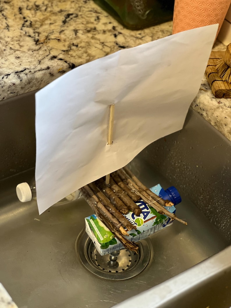 May include: A homemade boat made from a cardboard juice box, sticks, and a piece of paper for a sail. The boat is floating in a stainless steel kitchen sink.