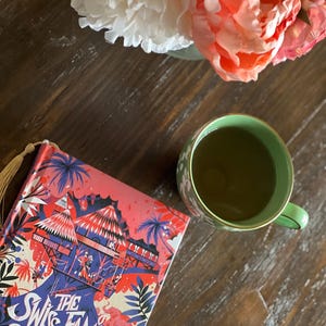 May include: A book titled "The Swiss Family Robinson" with a colorful illustration of a treehouse on the cover. A cup of green tea sits on a wooden table next to the book. Pink and white flowers are in the background.