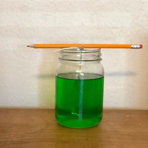 May include: A clear glass jar with a green liquid inside. A pencil is balanced on the top of the jar, with a string tied around the pencil and the jar.