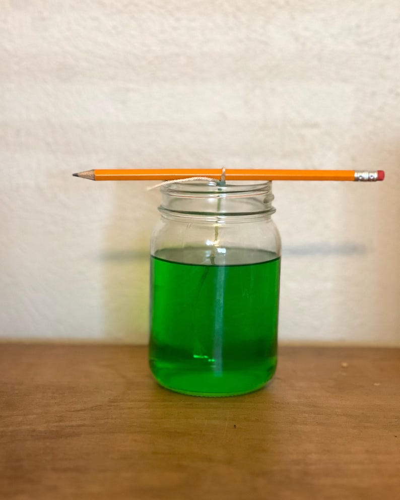 May include: A clear glass jar with a green liquid inside. A pencil is balanced on the top of the jar, with a string tied around the pencil and the jar.