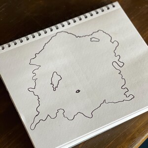 May include: A hand-drawn outline of an island with a smaller island inside it, drawn on a white sheet of paper in a spiral bound notebook.