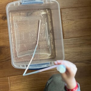 May include: A clear plastic container filled with water and a pink plastic straw. A hand is holding the straw above the container.