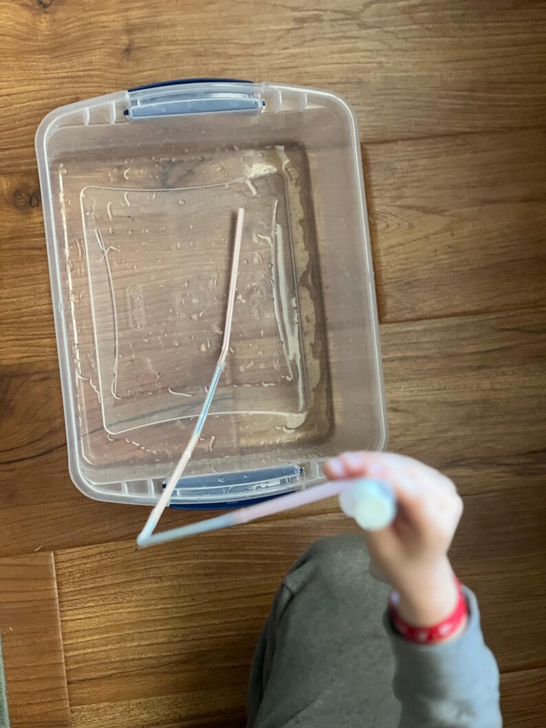 May include: A clear plastic container filled with water and a pink plastic straw. A hand is holding the straw above the container.