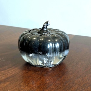 Puede incluir: Una calabaza decorativa de cristal con acabado negro y plateado. La calabaza tiene un tallo y está sobre una superficie de madera. Es un artículo de temporada, perfecto para la decoración de otoño o Halloween.