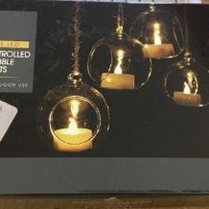 Puede incluir: Una caja negra que presenta seis luces de té de burbujas de vidrio con luces LED blancas cálidas. La caja incluye un mando a distancia y el texto "Remote Controlled Glass Bubble Tea Lights". La marca "NOMA SEASONS" es visible.