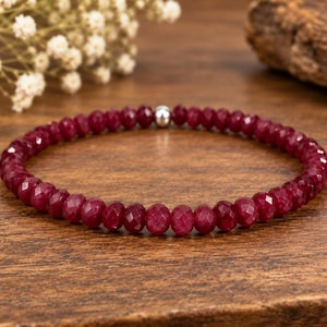 May include: A single-strand bracelet composed of small, faceted, deep red gemstone beads. The circular arrangement features a singular, polished silver-toned metallic bead positioned at the top centre, providing a subtle contrast to the uniform ruby-coloured stones.