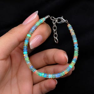 Puede incluir: Una pulsera compuesta por pequeñas cuentas circulares de ópalo iridiscente que muestran destellos de luz azul, verde y naranja. La pulsera cuenta con un cierre de mosquetón de metal plateado y una cadena extensora ajustable.