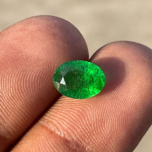 May include: A faceted, oval-shaped emerald gemstone. The gem is a vibrant green color and has a polished surface. The emerald is held between two fingers, showcasing its clarity and brilliance.