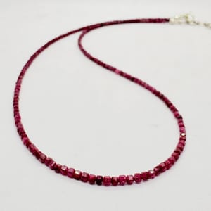 May include: A delicate necklace composed of small, faceted, deep magenta-red gemstone beads. The strand features a metallic silver-toned clasp mechanism at the end, providing a secure closure for this simple and refined piece of jewellery.