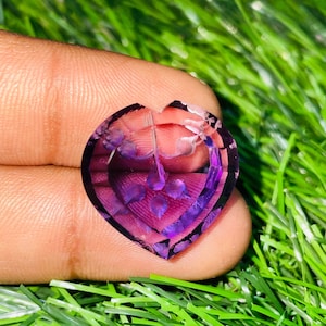 Puede incluir: Una piedra preciosa en forma de corazón con un diseño floral tallado. La gema presenta tonos de púrpura y violeta, con una superficie clara y pulida. La piedra preciosa se sostiene sobre un fondo de hierba verde.