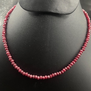 Puede incluir: Un collar compuesto por pequeñas piedras preciosas facetadas de color rojo intenso. Las cuentas están ensartadas para formar un círculo completo, exhibido sobre un fondo negro. El collar es una joya.