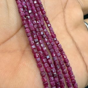 May include: A strand of small, square-cut, deep magenta-colored ruby beads. The beads are faceted, creating a reflective surface, and are strung together in a single, continuous line, showcasing a consistent size and vibrant, saturated red-pink hue.