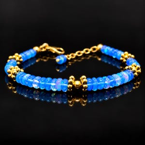 May include: A bracelet featuring round, translucent blue beads with iridescent highlights, interspersed with small gold-toned metal spacers and decorative clusters of tiny gold-toned spheres. The piece includes a gold-toned metal lobster clasp.