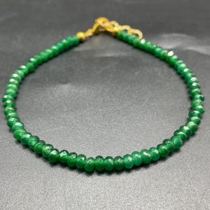 Puede incluir: Una pulsera delicada con pequeñas cuentas verdes facetadas. La pulsera tiene un cierre y una cadena dorados. Las cuentas son de un verde vibrante, y la pulsera se muestra sobre un fondo oscuro.