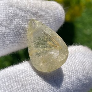 May include: A pear-shaped rutilated quartz gemstone featuring a clear, transparent body with visible golden, needle-like inclusions throughout the stone. The gemstone has a faceted surface that reflects light, showcasing the internal golden rutile strands.