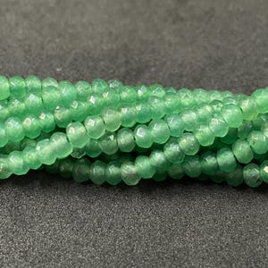 Może przedstawiać: Zbliżenie na pasma fasetowanych, jasnozielonych koralików. Koraliki mają lekko przezroczysty wygląd i polerowane wykończenie. Koraliki są nanizane razem, tworząc teksturowany i atrakcyjny wizualnie wyświetlacz. Tło to ciemna, teksturowana powierzchnia.