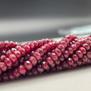 Könnte beinhalten: Nahaufnahme von Strängen facettierter, tiefroter Perlen. Die Perlen haben eine vielseitige Oberfläche, die das Licht einfängt und einen funkelnden Effekt erzeugt. Die Perlen sind aufgefädelt, was darauf hindeutet, dass sie für die Schmuckherstellung oder das Basteln bestimmt sind.