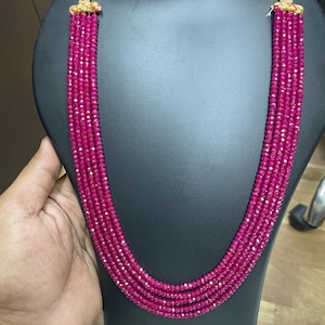 May include: A multi-strand necklace featuring five rows of small, faceted, vibrant magenta-pink beads. The strands are gathered at the top with gold-toned metal caps, creating a layered, cascading design that hangs in a deep U-shaped curve.