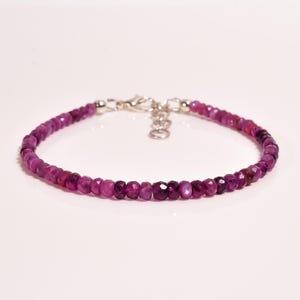 May include: A bracelet composed of small, faceted, deep magenta-coloured beads arranged in a continuous circular strand. The piece features a silver-toned metal lobster claw clasp and an adjustable extender chain with several circular links.