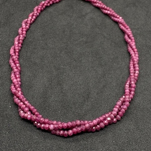May include: A multi-strand necklace composed of small, faceted, deep pink beads. The beads are arranged in a twisted, braided pattern, creating a textured, circular shape that highlights the reflective quality of the individual stone surfaces.