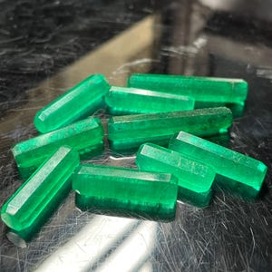 Op de afbeelding: Een verzameling levendige, smaragdgroene edelsteenkristallen. De kristallen zijn rechthoekig met facetranden, die het licht reflecteren. De edelstenen zijn verspreid over een reflecterend oppervlak.