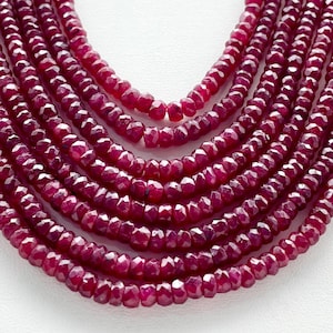 Puede incluir: Una hebra de cuentas de rubí, facetadas y pulidas, de color rojo intenso. Las cuentas están enhebradas en un collar de múltiples hebras, creando un aspecto lujoso y elegante.