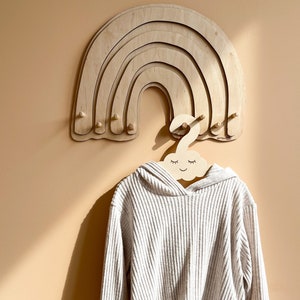 May include: A wooden rainbow wall hanger with five rounded arches and multiple hooks. A white hooded sweater hangs on a cloud-shaped hanger with a smiling face.